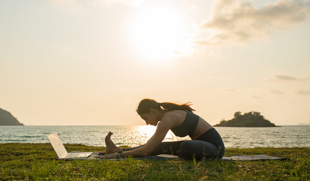 Young woman practicing yoga outdoors sea at sunset sitting on mat and performing online class on her laptop representing Seated Forward Fold – Paschimottanasana.